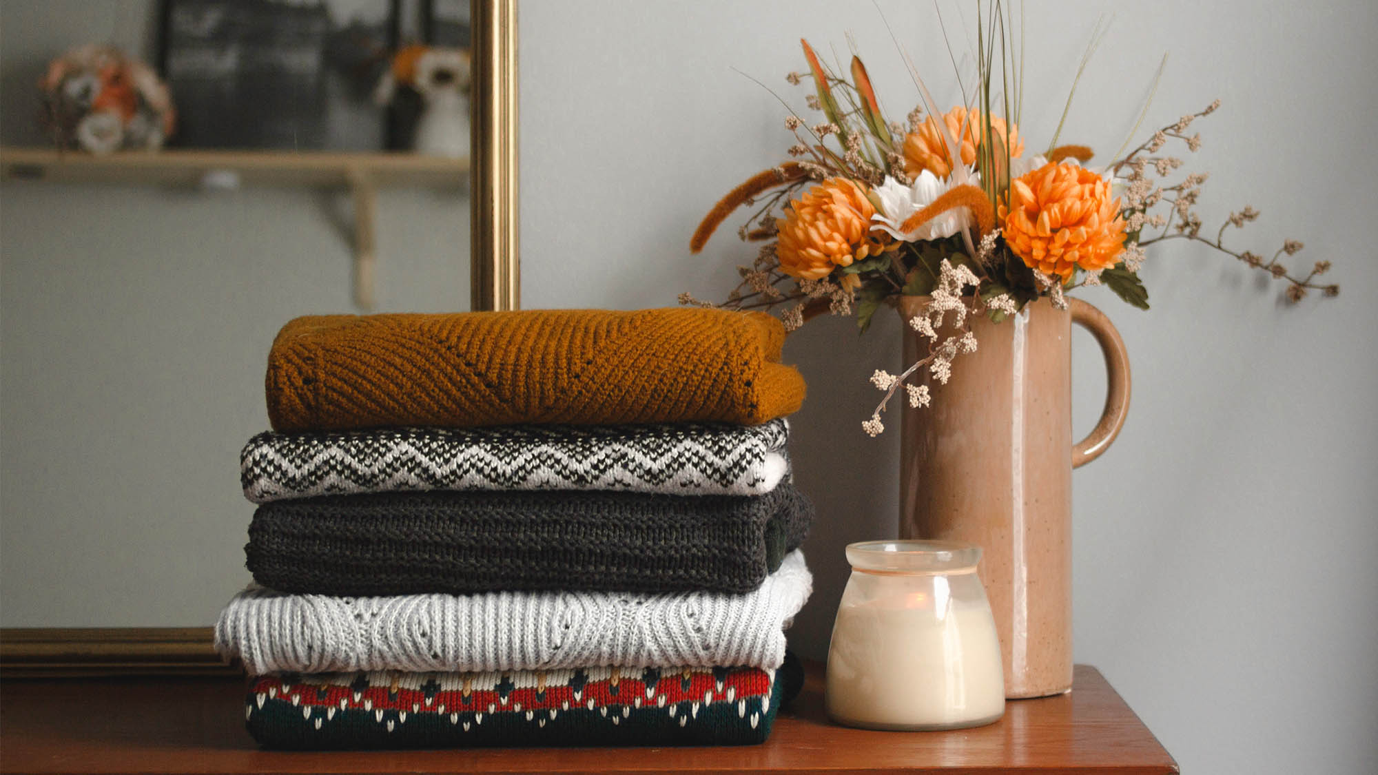 Five folded sweaters stacked on a dresser next to candle and vase with orange flowers