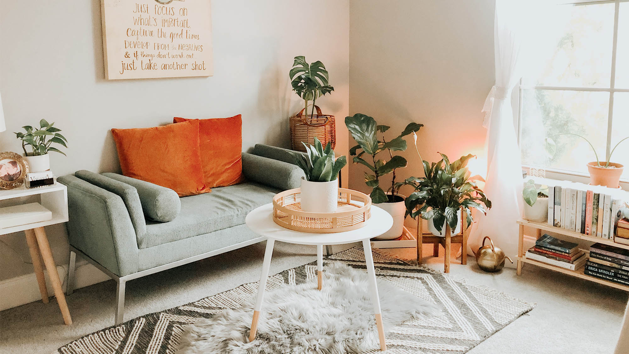 Sun-filled living room decorated with plants, gray couch and orange pillows, white table and small shag rug