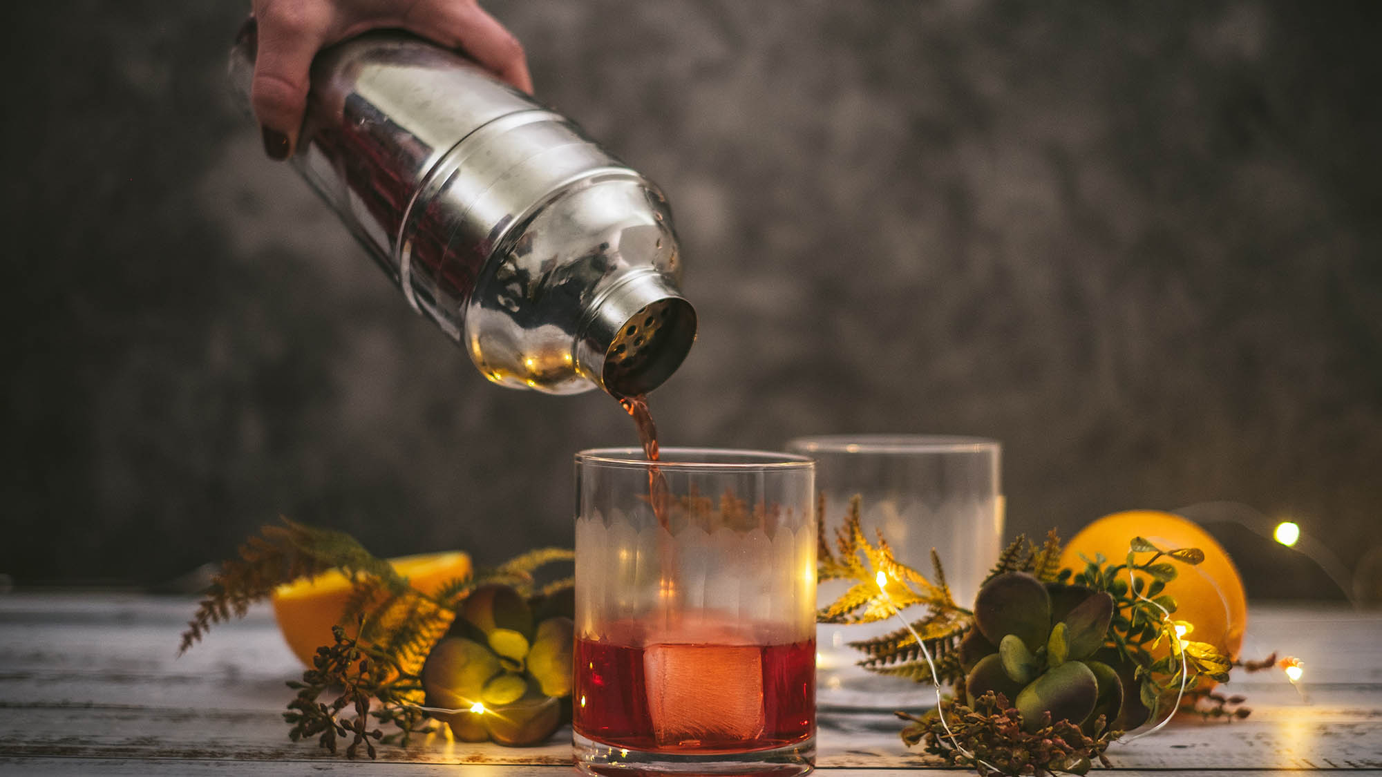 Negroni being poured from metal shaker into glass with ice cube amid sparkling lights and succulents