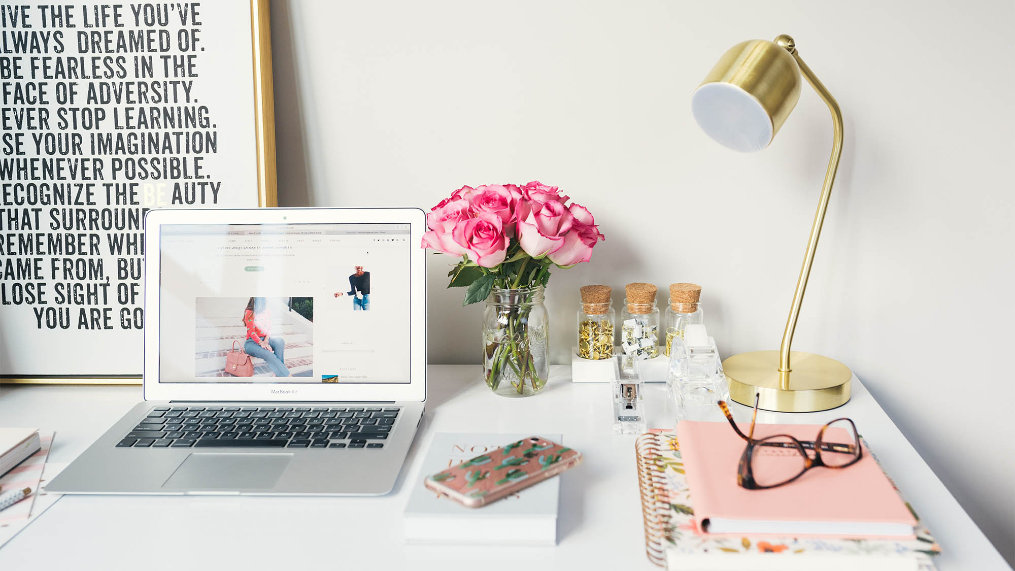 White desk with Macbook, pink roses, pink notepad and pink iPhone, framed art with words in background