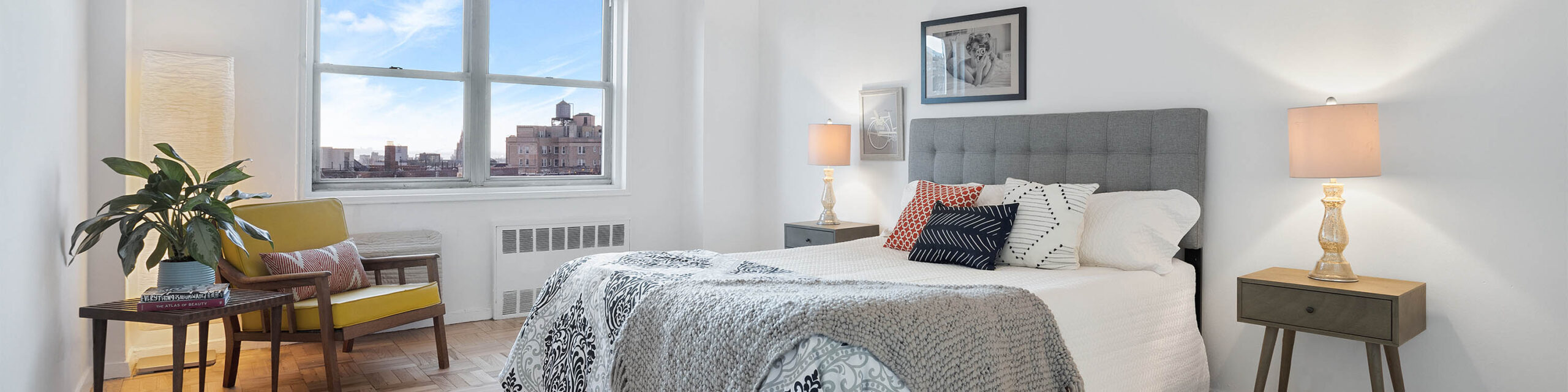 Staged bedroom with three multicolored pillows, gray headboard, two nightstands with lamps, wall art, and to the left two windows overlooking Brooklyn, a yellow chair and end table with plant