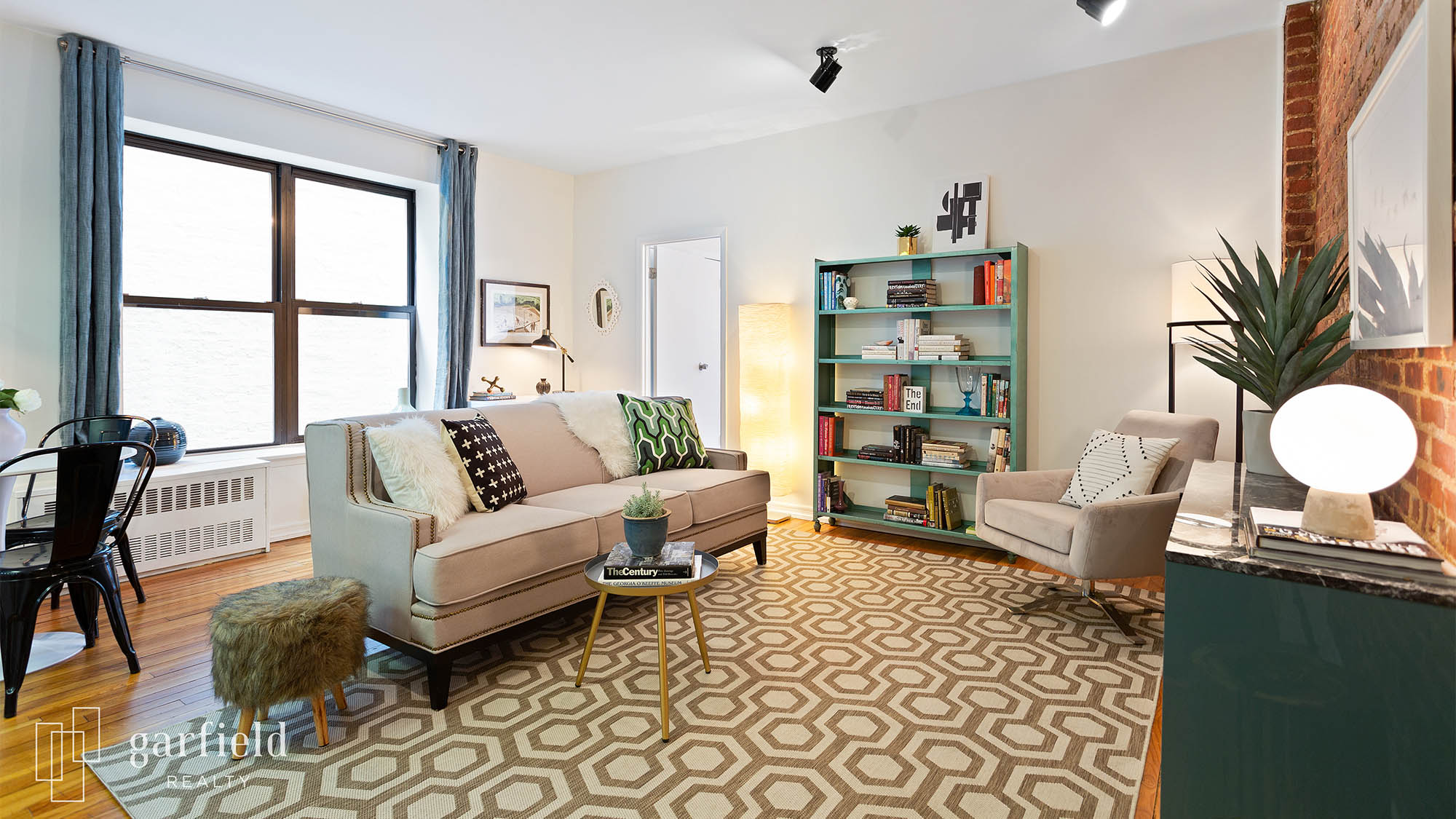 Staged living room featuring a couch with many pillows and a coffee table against a window with open blue curtain and a decorative shelf full of knick knacks and a chair on the right side