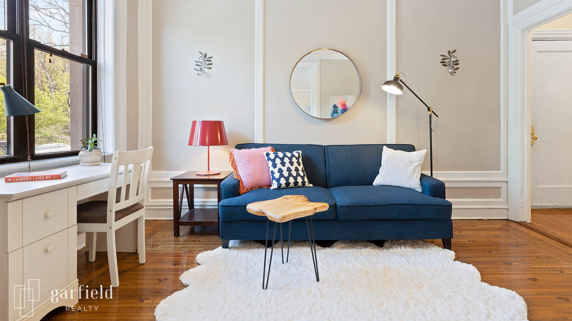 Staged living room with blue couch and modern decorative pillows with floor lamp to the right and coffee table to the left with red lamp and white desk and chair to the left against a window with white fur area rug and small wood table