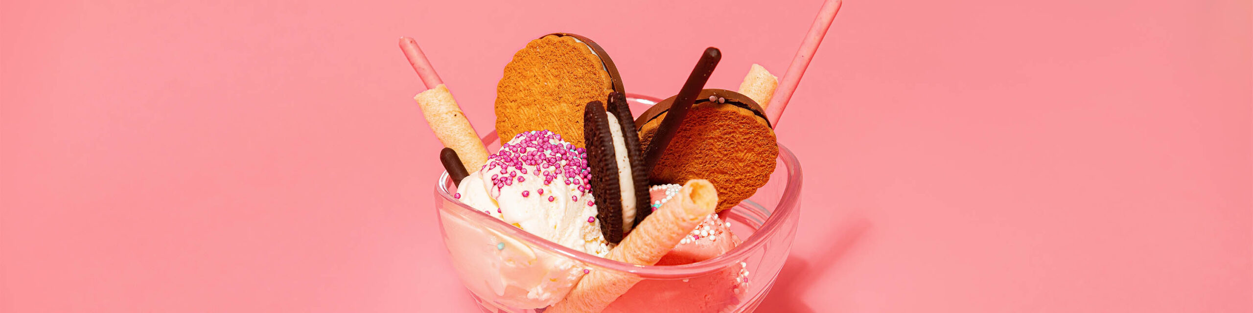Vanilla ice cream with pink sprinkles and cookies in clear glass dish on pink background