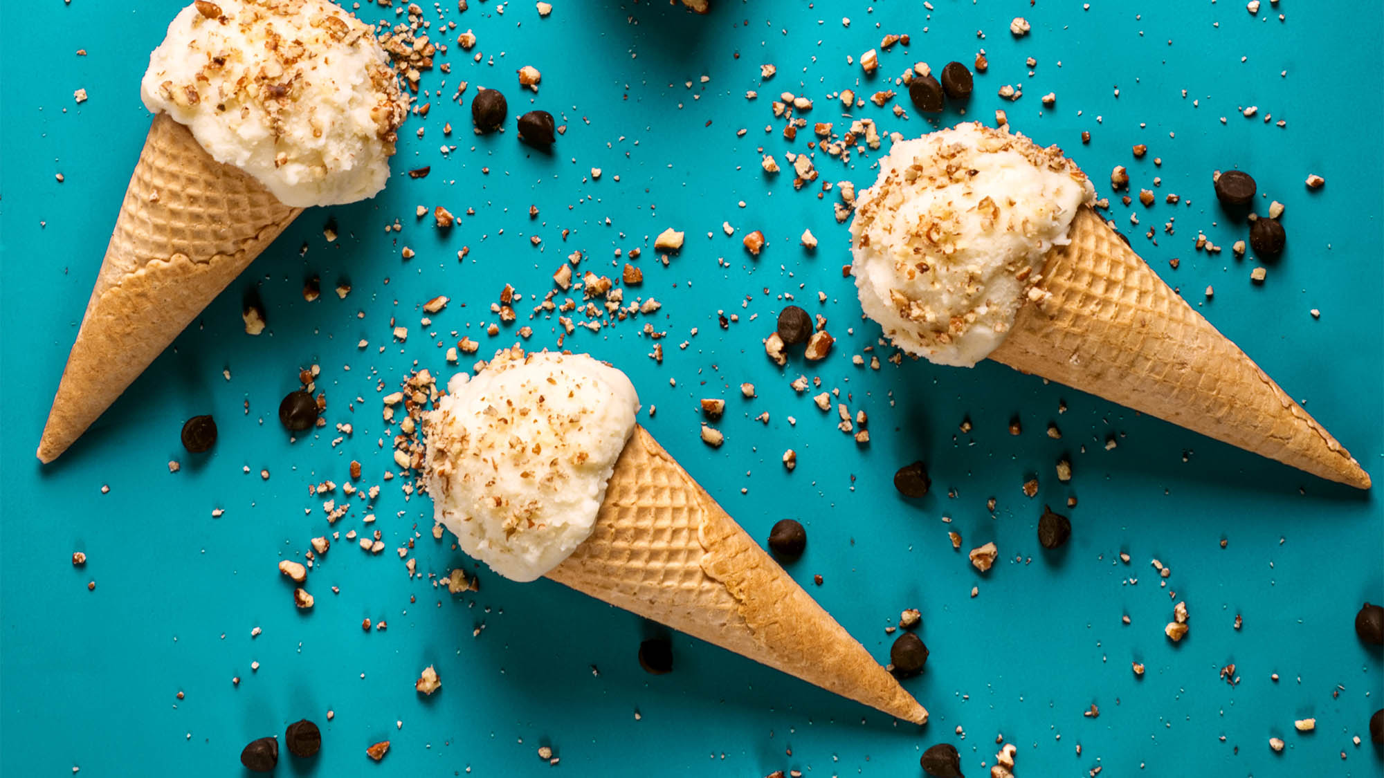 Three sugar cones with one scoop vanilla ice cream and chopped nuts resting on teal background with scattered crushed nuts