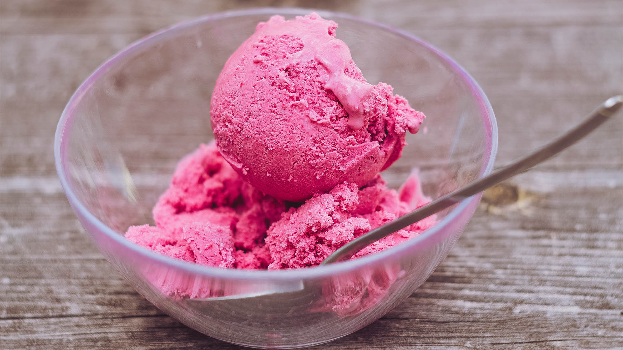 Clear glass dish of pink ice cream with spoon on top of wooden table