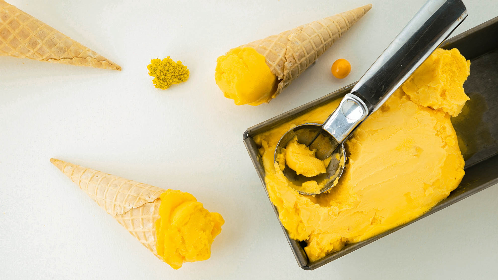 Two sugar cones with yellow ice cream and scoop in yellow ice cream container on white background