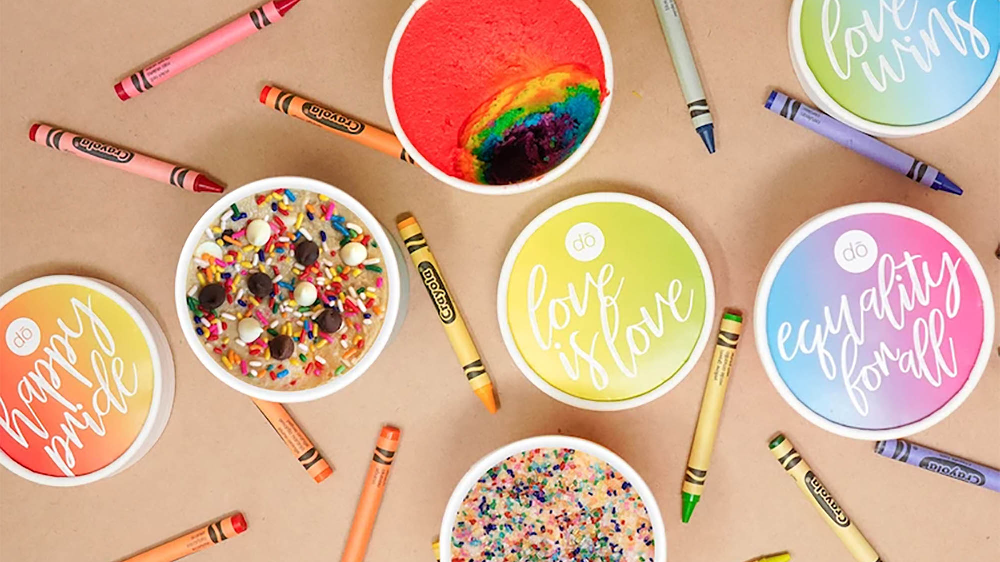 Rainbow edible cookie dough containers open and closed laid out with multicolored crayons on beige backdrop