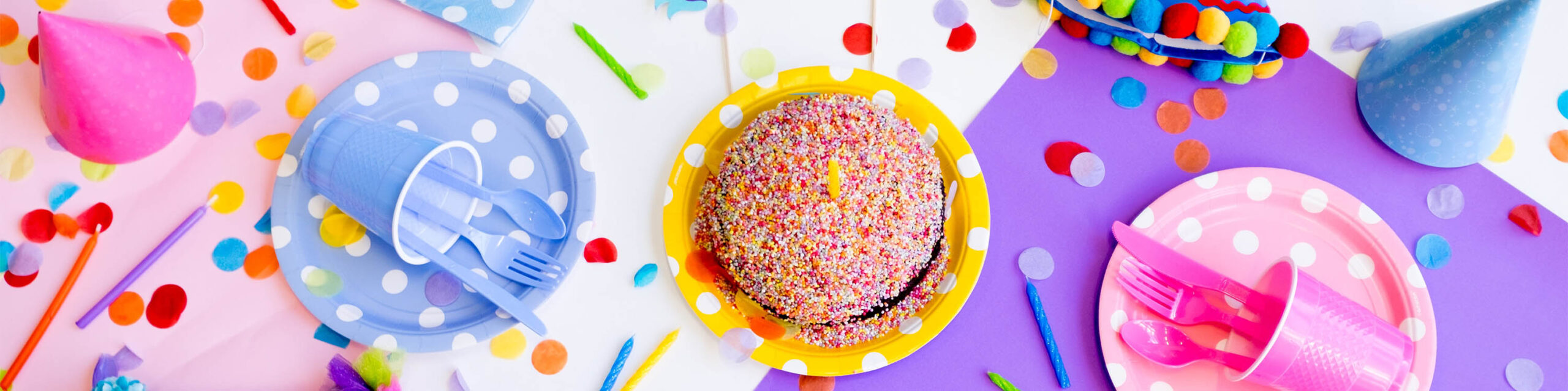 Lavendar plastic cup and utensils, rainbow sprinkle cake on yellow plate and pink plastic cup and utensils on party table with confetti decor