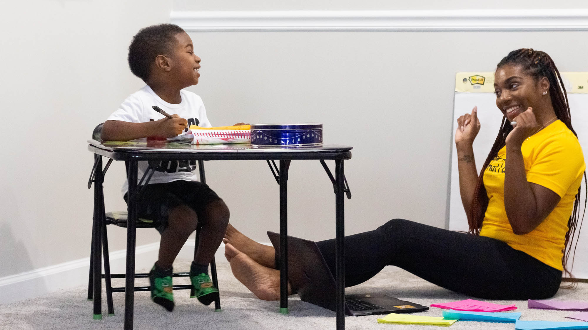 Black boy at table looking down at female Black teacher sitting on floor