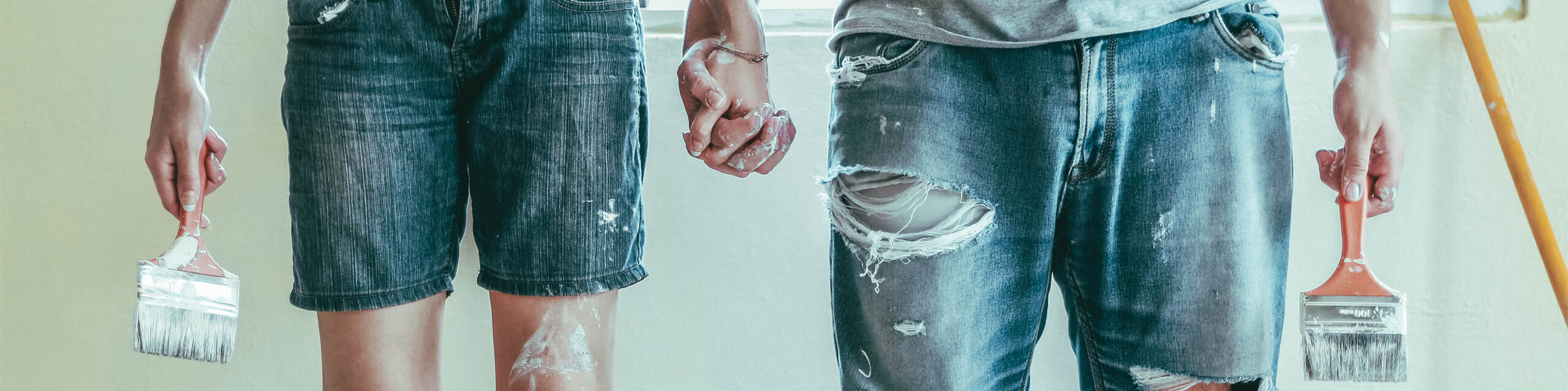 Female and male hips and hands closeup wearing denim and holding paintbrushes while holding hands