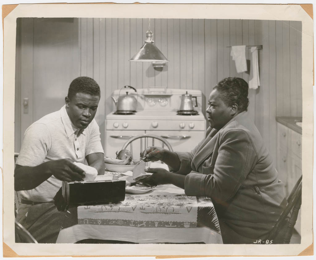 Black and white photo film still from Jackie Robinson movie with Jackie and Louise Beavers sitting at kitchen table with stove in background