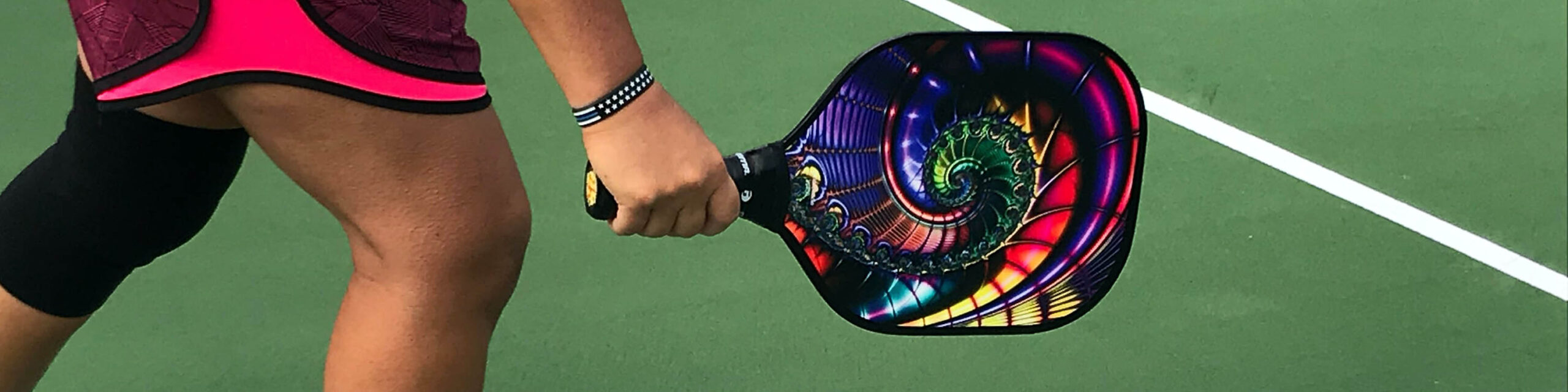 Persons knees and wrist holding rainbow pickleball racket on court