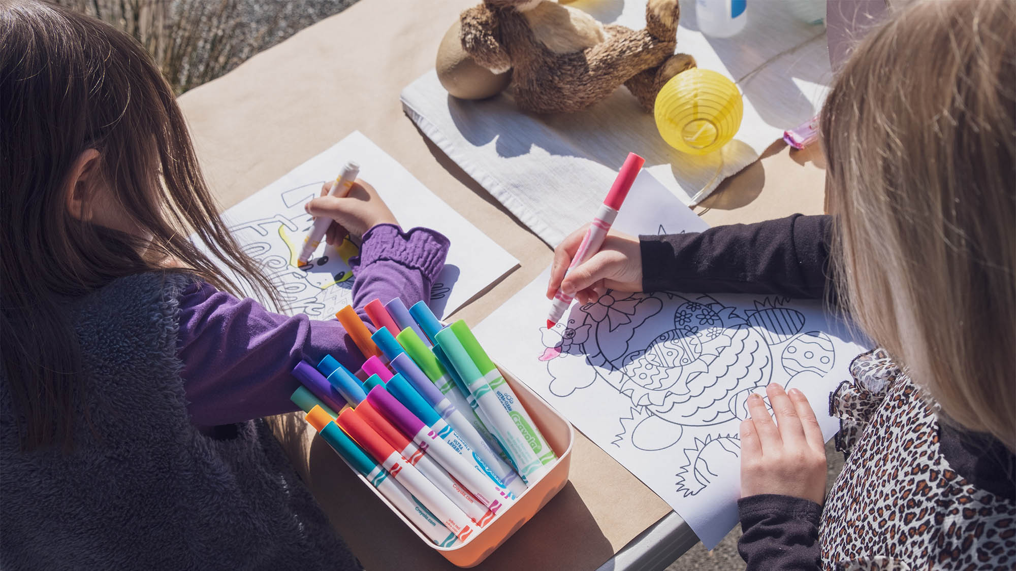 Overhead shot of two white girls sitting at outdoor table coloring Easter pictures with markers