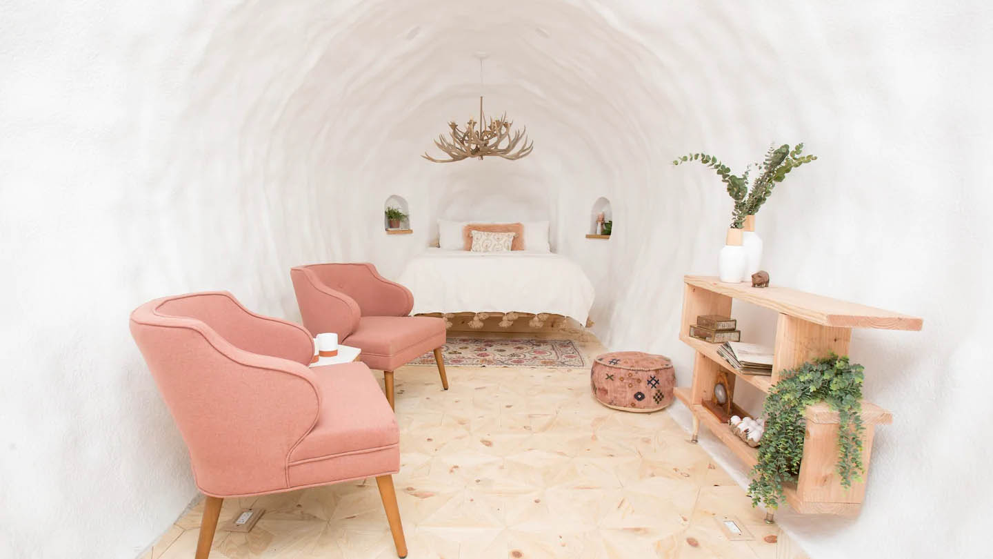 White walled hotel interior with two pink chairs on left and plant shelf on right and white bed in background with chandelier hanging above it