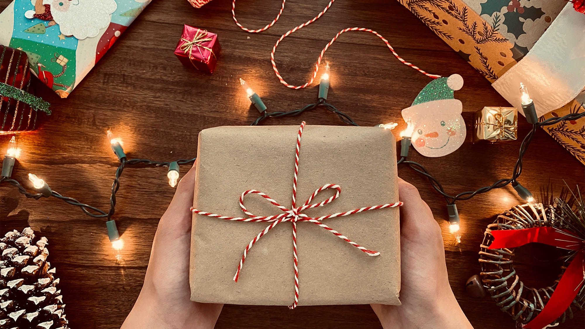 White hands holding brown wrapped package with red-white string above table with scattered Christmas ornaments and lights