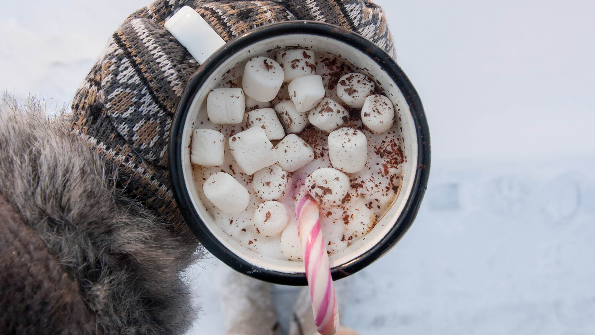 Mitten covered hand holding hot chocolate with marshmallows and candy cane