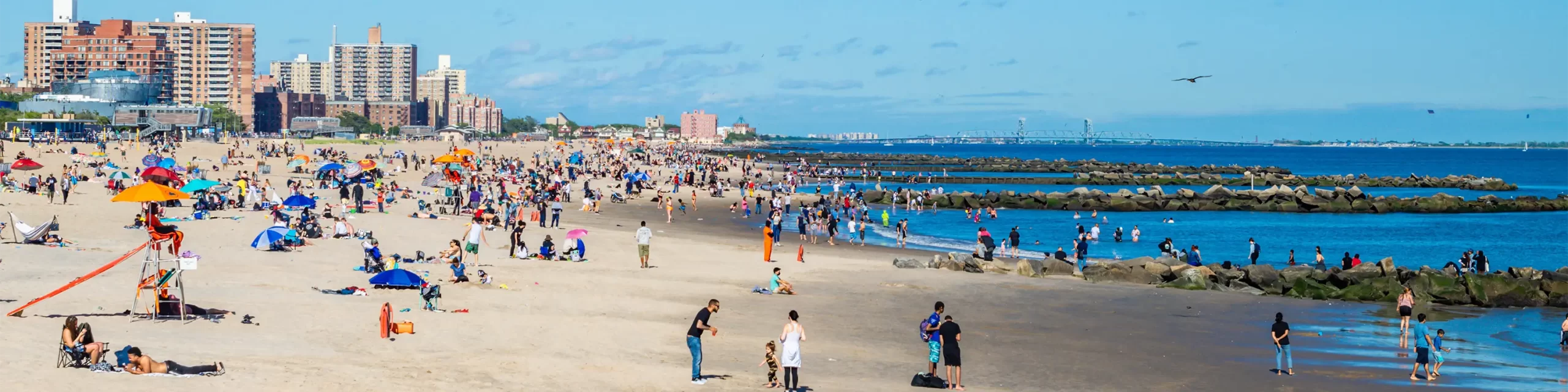 People on crowded Coney Island beach with tall buildings and water in background