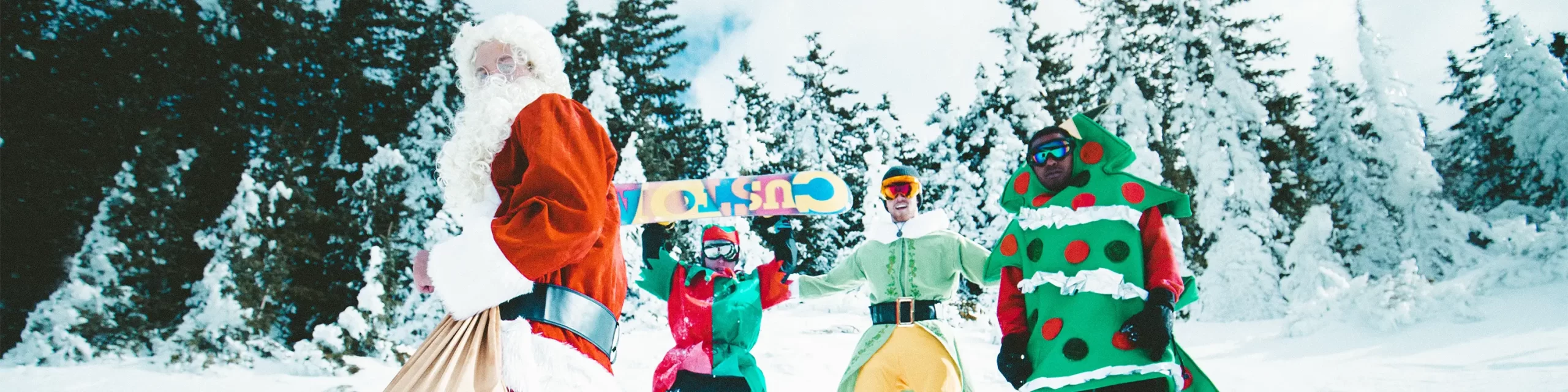 Snowboarders on snowy mountain dressed as Christmas elves, Santa and a Christmas tree