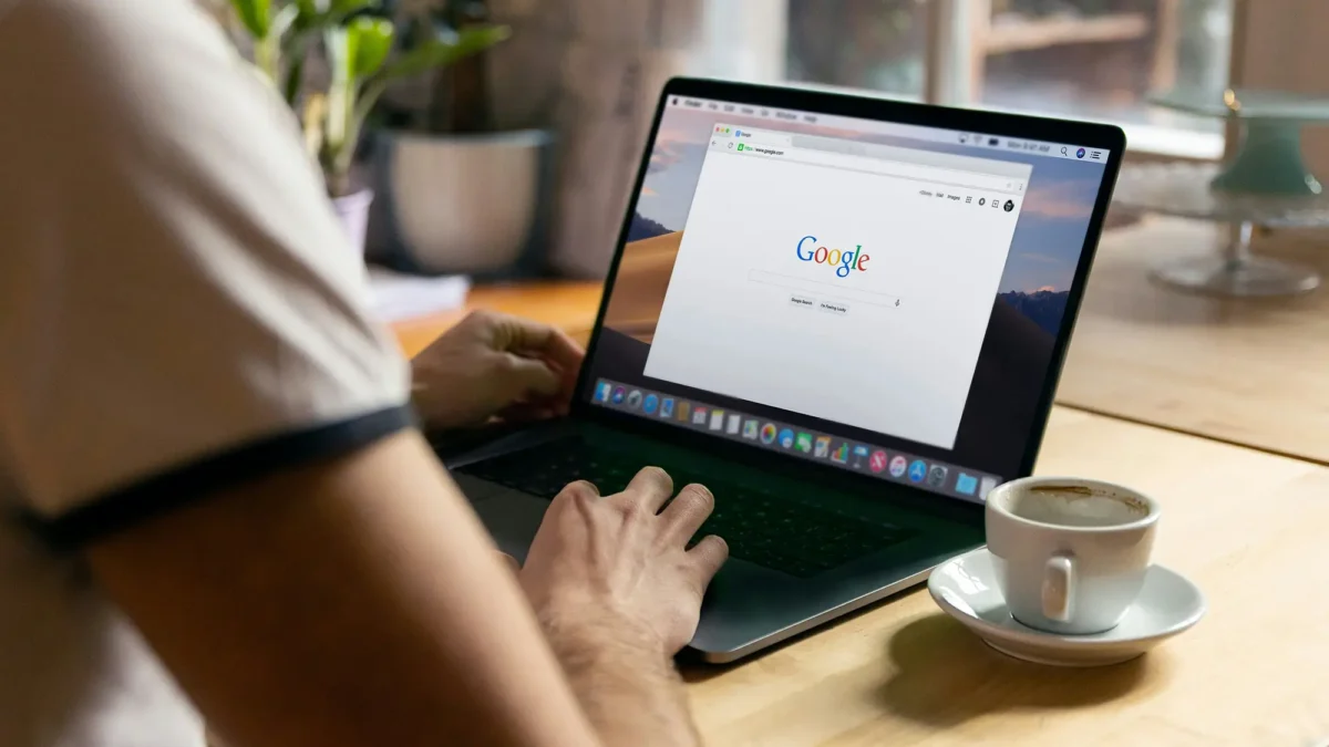 Persons hand on laptop keyboard with Google home screen and coffee cup and saucer beside it