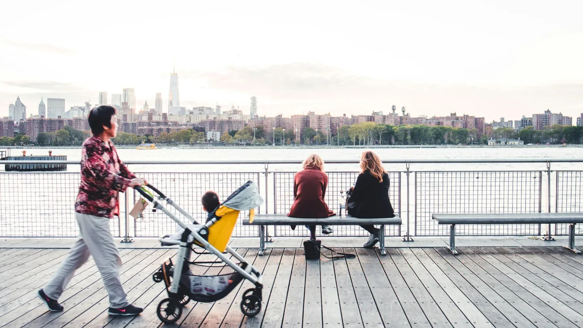 Woman pushing baby stroller and two women in background looking out at East River and NYC skyline