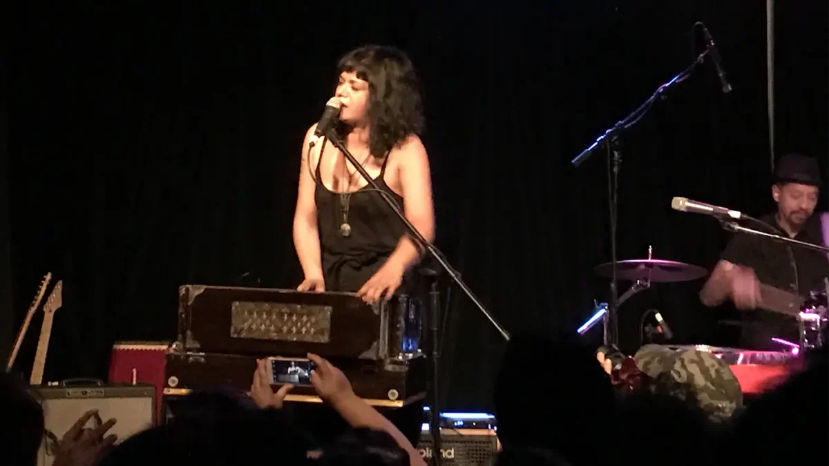 Woman on stage singing into microphone while playing harmonium