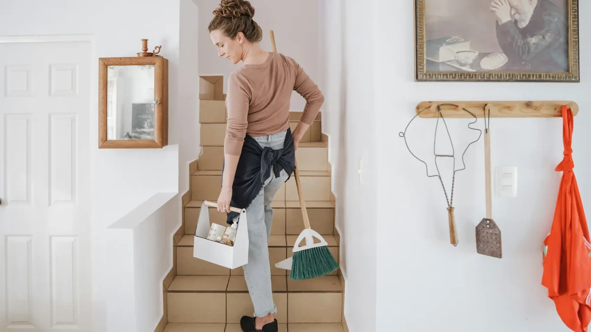 White woman holding cleaning products while walking up house stairs