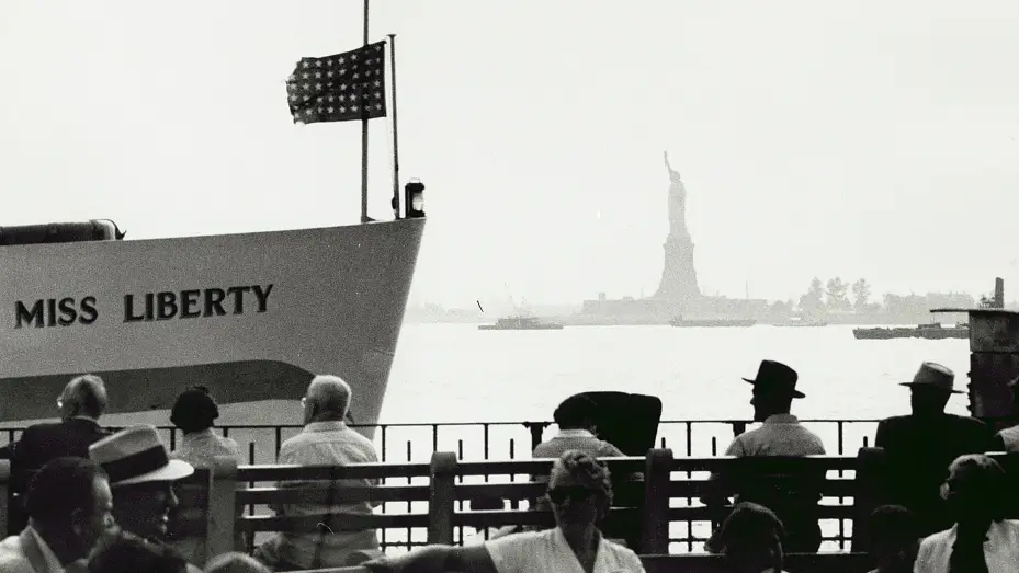 People sitting on dock overlooking Statue of Liberty and Miss Liberty Boat from 1954