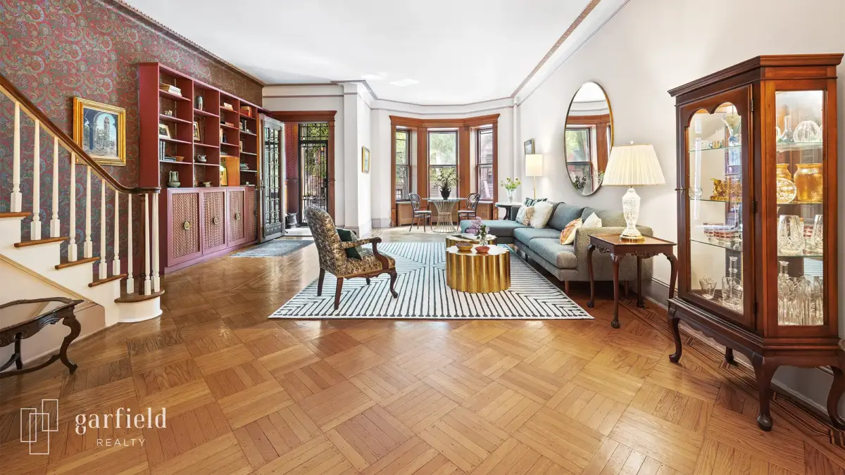 Living room with wood flooring, stair case on left, large area rug, chair on right and bay windows in rear